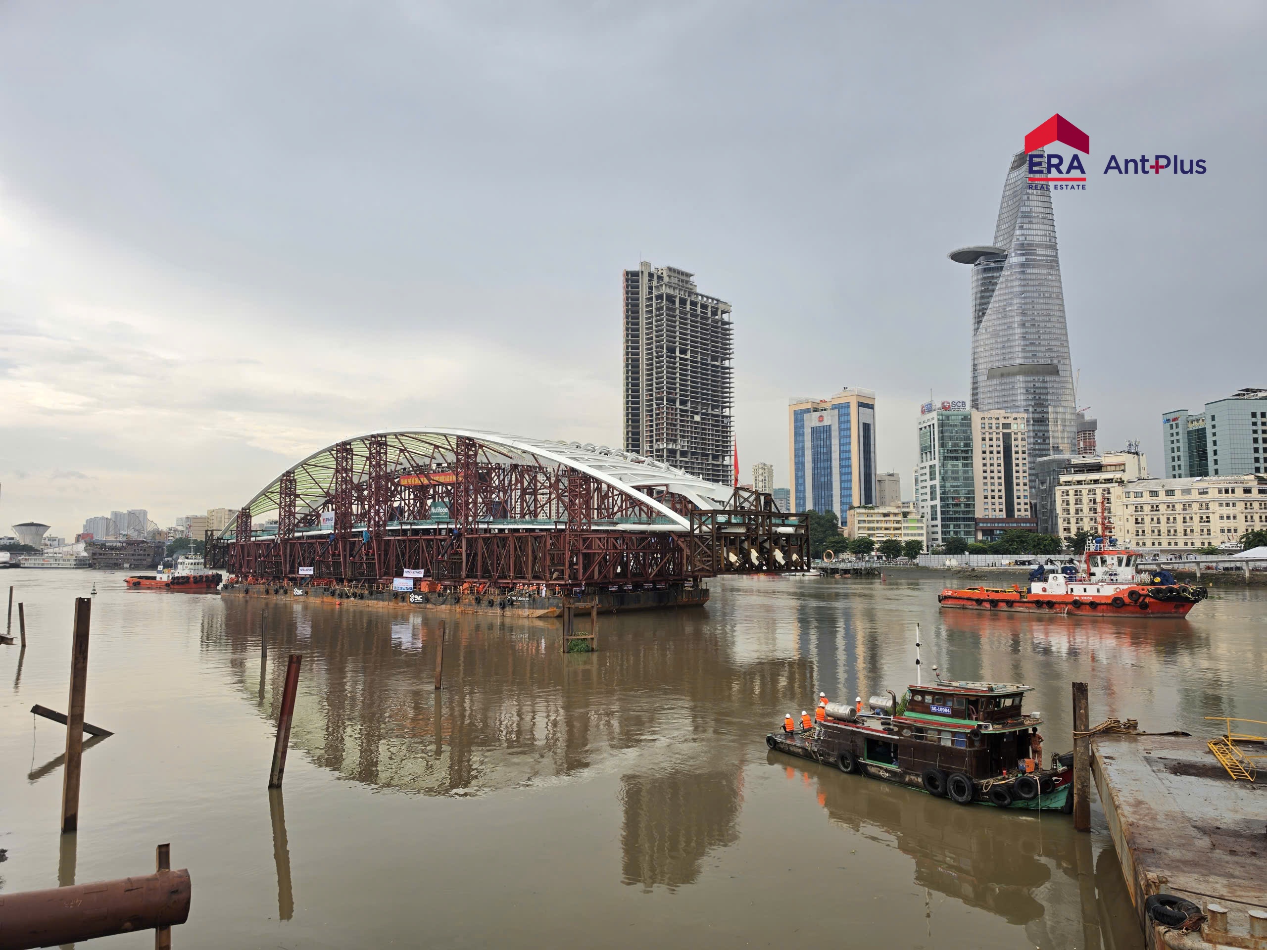 Sà lan chở nhịp chính cầu đi bộ Thủ Thiêm trên sông Sài Gòn cùng các tàu kéo màu cam, phía sau là tòa Bitexco